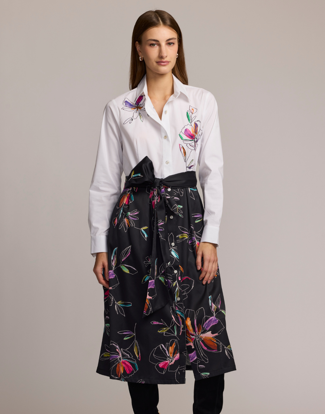 Woman wearing a shirt dress with a white floral embroidered top and black floral patterned skirt against a plain background. 