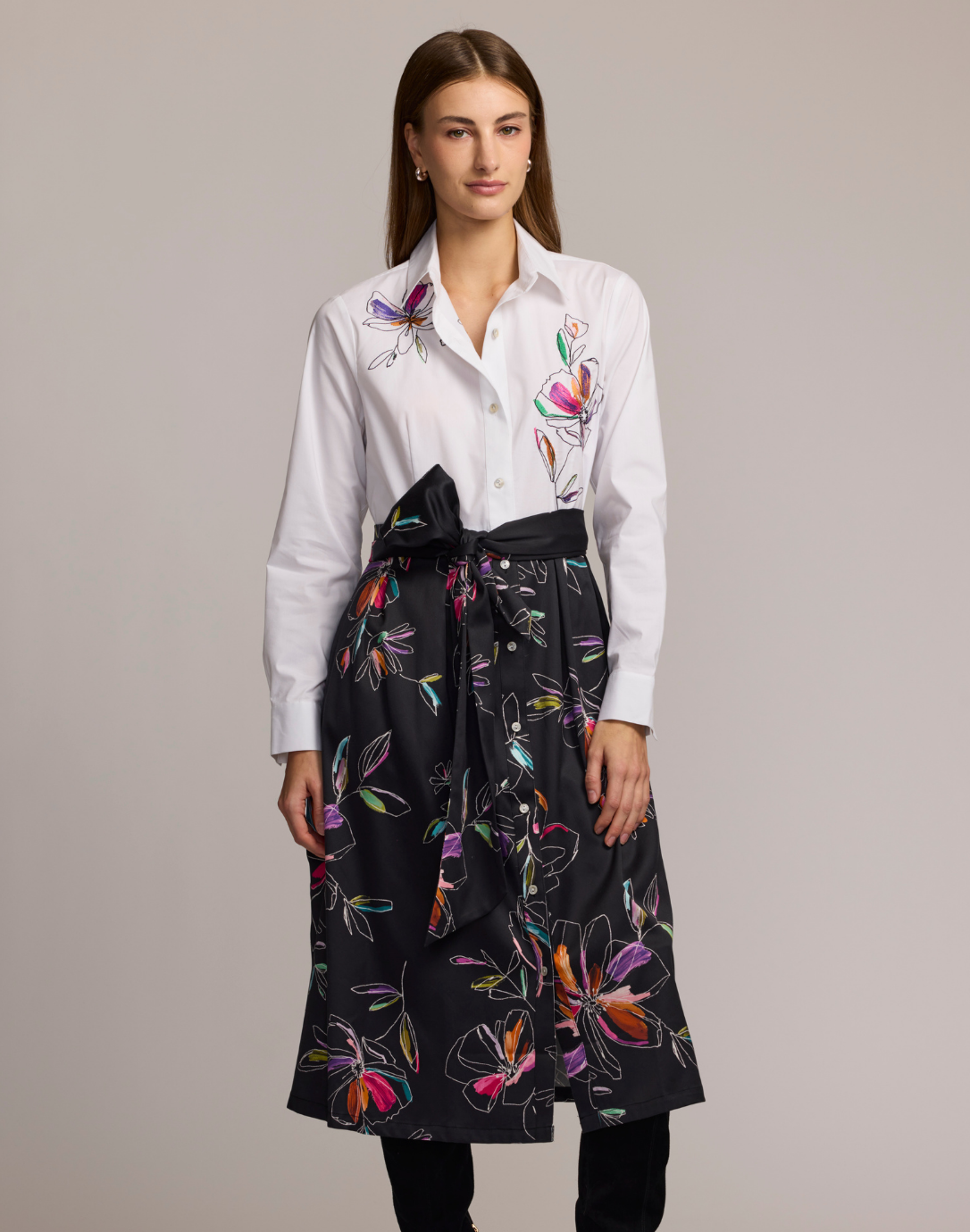 Woman wearing a shirt dress with a white floral embroidered top and black floral patterned skirt against a plain background. 