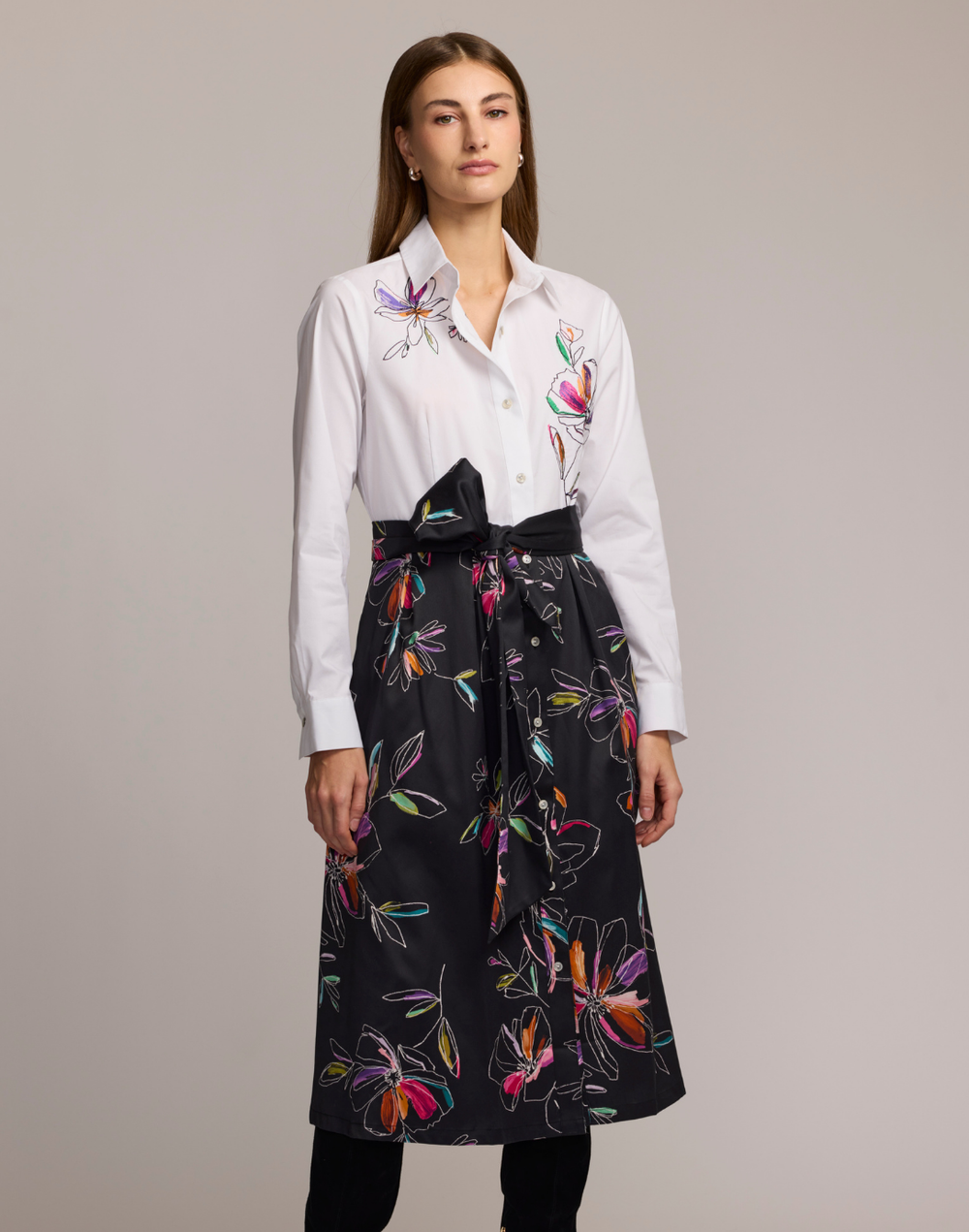 Woman wearing a shirt dress with a white floral embroidered top and black floral patterned skirt against a plain background. 