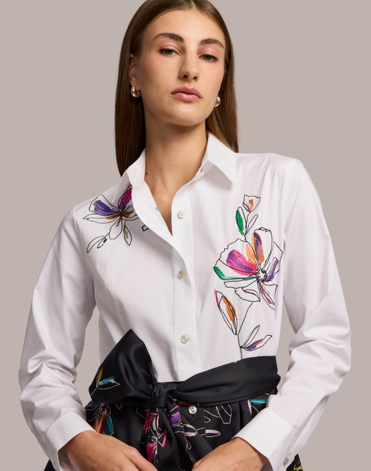 Woman wearing a shirt dress with a white floral embroidered top and black floral patterned skirt against a plain background. 
