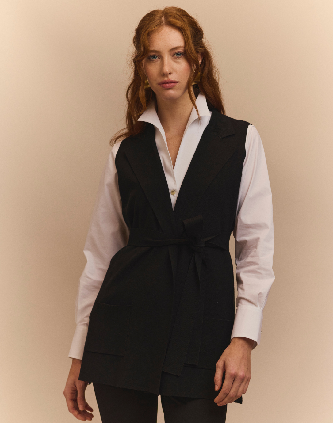 Woman wearing a black vest over a white shirt against a beige background